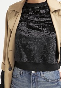 Černý sametový top s texturovaným povrchem a nařasenými detaily, kombinovaný s béžovým trenčkotem a modrými džínami.