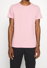 Camiseta de manga corta en suave algodón rosa claro con cuello redondo. Presenta un logo bordado sutil en el pecho. Textura suave.