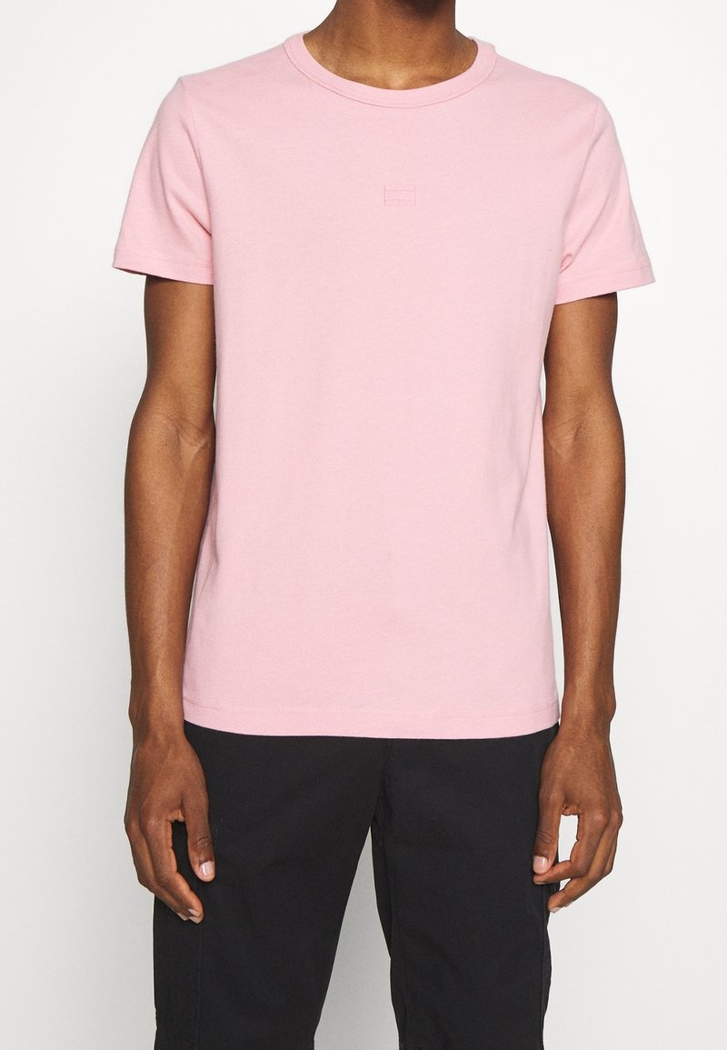 Camiseta de manga corta en suave algodón rosa claro con cuello redondo. Presenta un logo bordado sutil en el pecho. Textura suave.