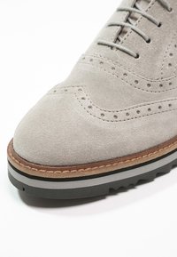 Šedé semišové brogue boty s dekorativními perforacemi, zaoblenou špičkou a vrstvovou černou a šedou gumovou podrážkou s hnědým koženým lemem.