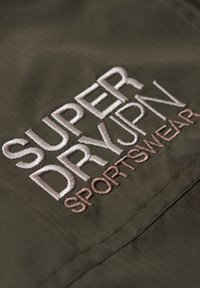 Olivegrøn stof med et rutenetmønster; har broderet "SUPERDRY" i hvid og "SPORTSWEAR" i lysebrun.