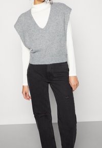 Grå stickad tröja med v-ringning och korta ärmar bärs över en vit långärmad topp, ihop med svarta slitna jeans.