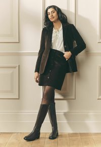 Barna kordbársony blézer és szoknya szett, gombos részletekkel, barna térdig érő bőr csizmával és fehér, csipkés díszítésű blúzzal párosítva.