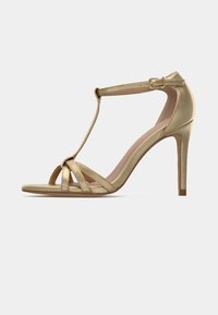 Kulta korkokenkiä T-nauhalla, nilkkahihnalla ja kolmella etuhihnalla beige pohjalla, esillä valkoista taustaa vasten.