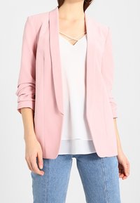 Světle růžový přizpůsobený blazer s dlouhými rukávy, se zářezovým límcem a otevřeným předním dílem, doplněný o bílé tričko a světle modré džíny.