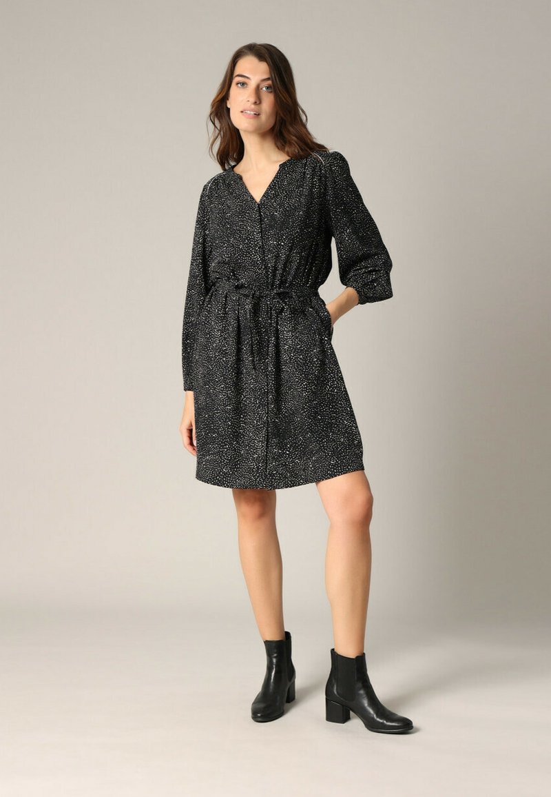Robe noire à motif avec des manches longues, taille cintrée et décolleté en V. Associée à des bottines noires pour le contraste. Texture de tissu lisse.