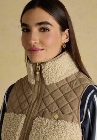 Kohverdatud tan värvi vest beezist karvast kraest ja rinna taskutega, millel on kuldne tõmblukk ja riistvara. Triibuline särk on osaliselt nähtav all.