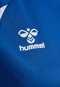 Logo Hummel avec abeille stylisée et texte sur tissu de sport bleu avec accents blancs, montrant la texture et les détails de la couture.