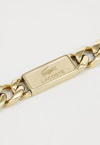 Bracelet en chaîne dorée présentant une plaque rectangulaire texturée avec le logo Lacoste gravé. Les maillons ont une finition polie, ajoutant une qualité réfléchissante.