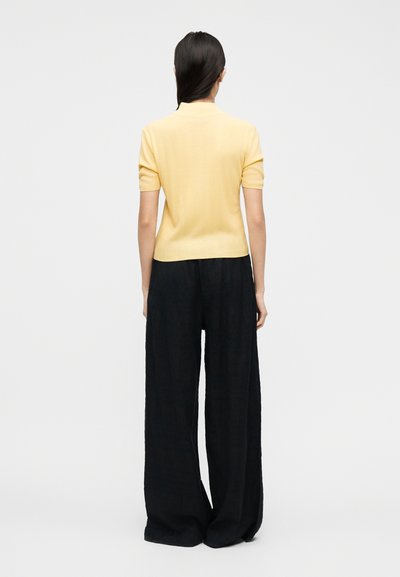 Γυναίκα που στέκεται με την πλάτη στην κάμερα, φοράει κίτρινο τοπ με κοντά μανίκια και φαρδιά μαύρη παντελόνα εναντίον απλού φωτεινού φόντου.