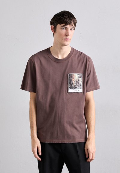 Barna pamut póló, mellzsebével, amely fekete-fehér városkép mintával díszített, kerek nyakkivágással és rövid ujjakkal.