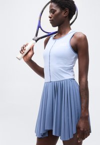 Γυναίκα με ανοιχτό μπλε φόρεμα τένις που κρατά ρακέτα τένις ακουμπισμένη στον ώμο της, με φόντο απλό.