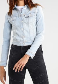 Hellblaue Jeansjacke mit figurbetontem Schnitt, vorderen Druckknöpfen, zwei Brusttaschen und kurzer Länge, kombiniert mit schwarzen, strukturierten Hosen.