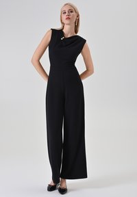 Combinaison noire avec un col drapé et des boutons dorés, caractérisée par des jambes larges et une texture de tissu lisse. Portée avec des ballerines noires.