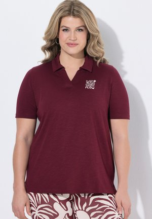 Kvinde med bølget blondt hår iført en vinrød kortærmet poloshirt med "FLOW"-logo og mønstrede vinrøde og hvide bukser, stående mod en hvid væg.
