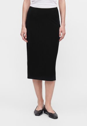 Femme portant une jupe crayon midi noire, un haut blanc et des chaussures plates noires, debout devant un fond blanc uni.