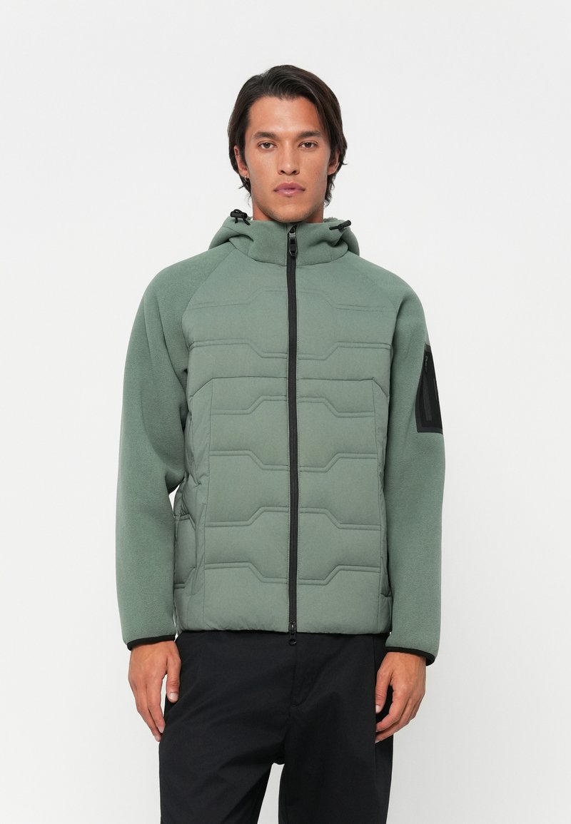 Veste en polaire verte avec fermeture éclair à l'avant, texture matelassée avec des motifs hexagonaux et accent noir sur la manche gauche.