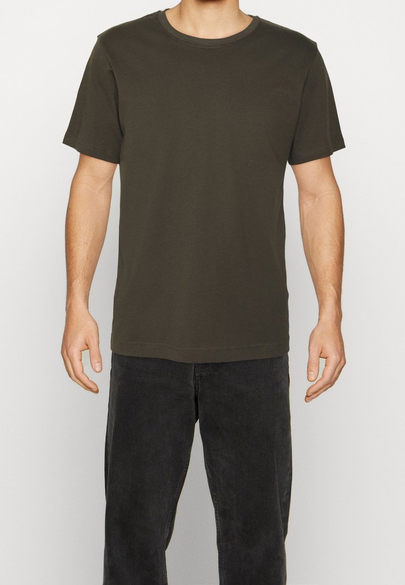 Homme portant un t-shirt uni vert olive foncé à manches courtes et col rond, ainsi qu’un jean noir, debout sur un fond blanc.