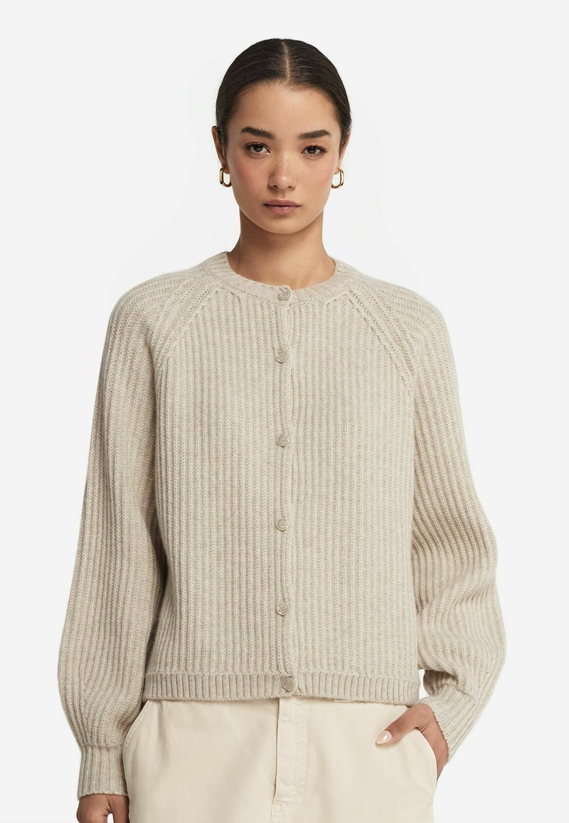 Cardigan beige à côtes avec un col rond, des boutons sur le devant et des manches longues. Le tissu a une texture douce avec des variations tonales subtiles.