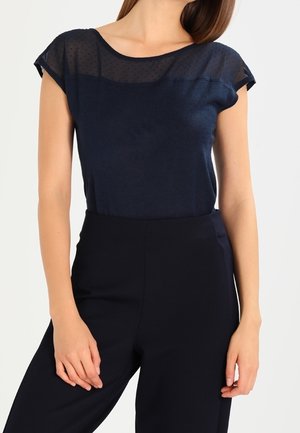 Marineblaues Top mit einem durchsichtigen, gepunkteten oberen Abschnitt und kurzen Ärmeln, kombiniert mit hochtaillierten dunklen Hosen. Weicher Stoff und lockere Passform.