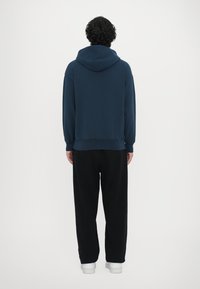 Námořnická mikina s kapucí a elastickými manžetami, v kombinaci s černými volnými kalhotami a bílými teniskami. Jednoduchý, minimalistický design.