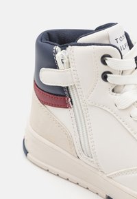 Sneaker alta bianca e blu navy con un accento rosso, caratterizzata da una tomaia in pelle liscia, camoscio testurizzato, dettaglio con zip e lacci bianchi.