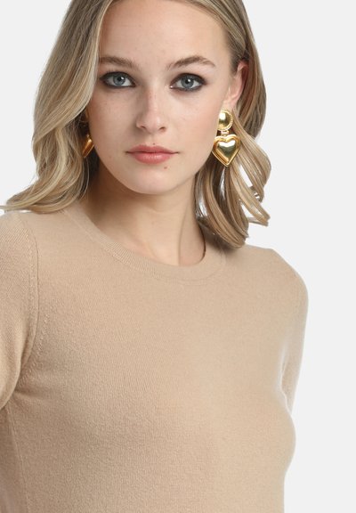 Bežs adīts džemperis ar apaļu kakla izgriezumu. Lieli zeltaini sirds formas auskari. Gluda tekstūra, smalka šuvju detaļa uz piedurknēm.