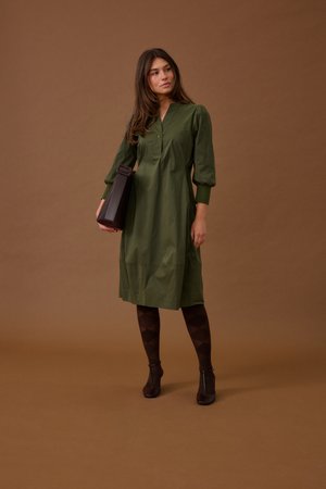 Nainen oliivinvihreässä, pitkähihaisessa midimekossa, kuviollisissa sukkahousuissa ja nilkkureissa, pitäen tummaa ruskeaa käsilaukkua ruskealla taustalla.