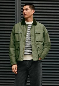 Olivgrüne Jacke mit Cordkragen, mehreren vorderen Taschen und Reißverschluss. Getragen über einem strukturierten schwarz-weißen Pullover und dunkelgrauen Hosen.