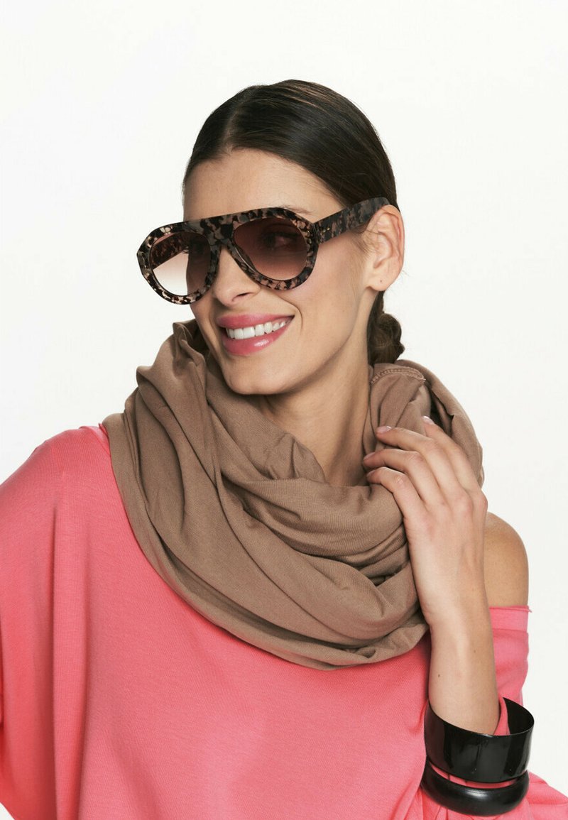 Brązowe, oversize'owe okulary przeciwsłoneczne z wzorzystą oprawką, zestawione z beżowym szalem i luźną, różową bluzką z odkrytymi ramionami.