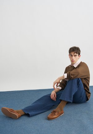 Giovane uomo seduto su un tappeto blu, indossa un maglione marrone, pantaloni blu, calzini marroni e scarpe di pelle marrone, su uno sfondo bianco uniforme.