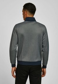 Man gezien van achteren, gekleed in een grijze jas met lange mouwen en een marineblauwe kraag en manchetten, gecombineerd met zwarte broek.