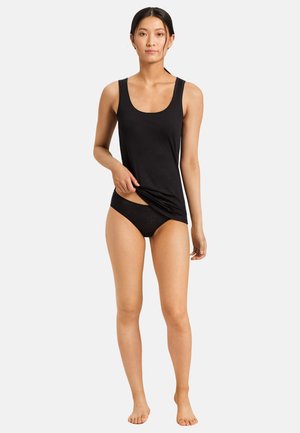 Kvinde med langt sort hår, der står barfodet iført sort tanktop og sort undertøj, løfter let kanten med den ene hånd.