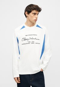 Νεαρός άνδρας που φοράει λευκό πουκάμισο με μακριά μανίκια με μπλε λεπτομέρειες και μαύρο χειρόγραφο κείμενο μπροστά, κοιτάζοντας προς τα δεξιά του.