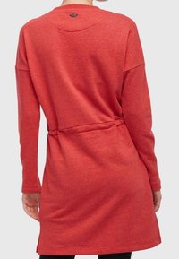Robe pull rouge avec des manches longues, une taille cintrée et un col rond. Texture douce avec un logo subtil au-dessus du col.