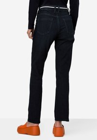 Jeans rectos de color azul oscuro con un cinturón y dos bolsillos traseros, combinados con zapatos de plataforma naranjas sobre un fondo claro.
