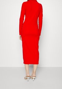 Robe ajustée à manches longues rouge, longueur midi, avec un léger col, fabriquée en tissu lisse. Associée à des talons blancs à lanières.
