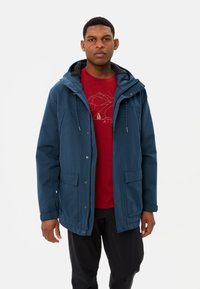 Giacca outdoor blu con cappuccio, due tasche frontali e chiusura a pressione. Indossata sopra una maglietta rossa con grafica raffigurante un design montano.