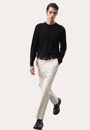 Mann, der geht, trägt ein schwarzes Langarmhemd, beige Hose, schwarzen Gürtel und schwarze elegante Schuhe vor einem schlichten hellen Hintergrund.