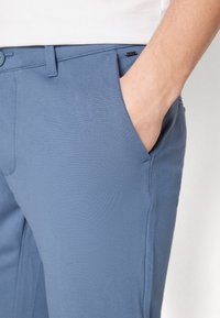 Modré kalhoty vyrobené z hladké tkaniny, se přední kapsou, poutky na opasek a zapínáním na knoflík. Boční šev viditelný pro přidaný detail.
