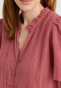 Rosa texturerad blus med vertikala ränder, volangkrage, knappstängning fram och korta puffärmar. Lätt tyg med subtila skimrande detaljer.