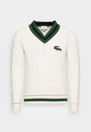 Biały sweter z prążkowanym splotem w serek, z ciemnozielonym i granatowym paskiem przy kołnierzu, zielonym i żółtym paskiem przy dolnej krawędzi oraz zielonym logo krokodyla na piersi.