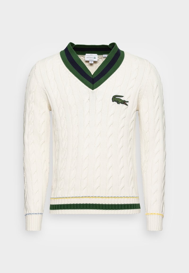 Pull en maille blanche à col en V avec rayures vert foncé et bleu marine sur le col, rayures vertes et jaunes à l'ourlet, et un logo crocodile vert sur la poitrine.