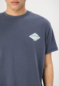 Camiseta de algodón azul marino con cuello redondo, que presenta un logo blanco en forma de diamante con el texto "TOMMY JEANS" y el año de establecimiento de 1985.