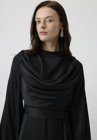 Must mustriga mustan riidega, millel on kumer kaelus, pikad varrukad ja satiinimaal. Omab struktureeritud talje, millel on sobiv vöö. Elegantne ja voolav kujundus.