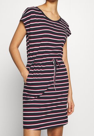 Kobieta ubrana w granatową sukienkę z białymi i czerwonymi poziomymi paskami, krótkimi rękawami i ściągaczem w talii, stoi z jedną ręką w kieszeni.