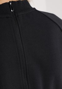 Czarny top sportowy z wysokim kołnierzem, centralnym zamkiem błyskawicznym i gładką teksturą materiału, o eleganckim, minimalistycznym designie.