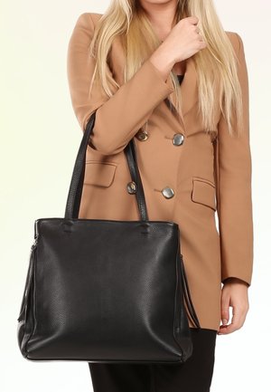 Femme en blazer beige tenant un grand sac à main en cuir noir avec doubles anses sur fond uni.