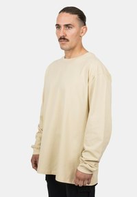 Beige Baumwollhemd mit langen Ärmeln, rundem Halsauschnitt, lockerer Passform und abfallenden Schultern, präsentiert an einem männlichen Modell vor einem weißen Hintergrund.