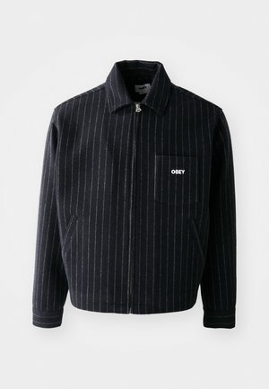 Chaqueta azul marino con rayas blancas verticales, cierre frontal con cremallera, cuello con solapa, bolsillo en el pecho y logo "OBEY" en blanco.